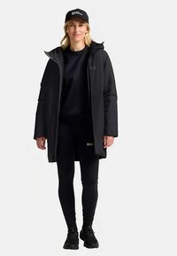 Schwarzer langer Mantel mit Kapuze, Reißverschluss vorne und weichem Futter über einem schwarzen Sweatshirt und Leggings. Schwarze Sneaker und eine Mütze vervollständigen das Outfit.