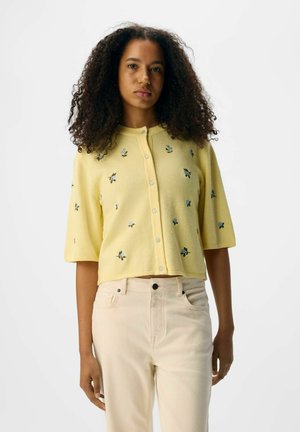 Gul kortärmad cardigan med korta ärmar, prydd med blå blommig broderi, knappstängning och en mjuk stickad textur. Matchad med ljusa jeans.