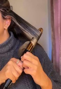 Un fer à friser noir avec des accents dorés boucle des cheveux châtains clairs. Des mains s'agrippent à l'appareil tout en portant une bague en argent. Un pull en col roulé gris est visible.