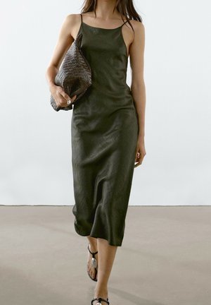 Donna che cammina indossando un vestito sottoveste verde scuro e sandali neri, tenendo una grande pochette intrecciata marrone scuro.