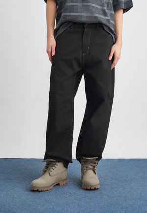 Pantalones de mezclilla negra con costuras amarillas en contraste, de corte recto. Combinados con botas de ante gris con suela de goma y diseño de cordones.