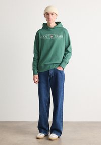 Felpa verde in cotone con grafica frontale, abbinata a jeans blu a vestibilità rilassata e sneakers bianche con suole beige. Berretto lavorato a maglia semplice.