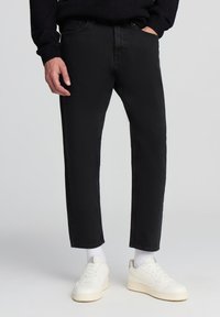 Jeans in denim nero con taglio dritto, due tasche frontali e chiusura con zip. Abbinati a scarpe da ginnastica bianche e calzini bianchi alla caviglia.