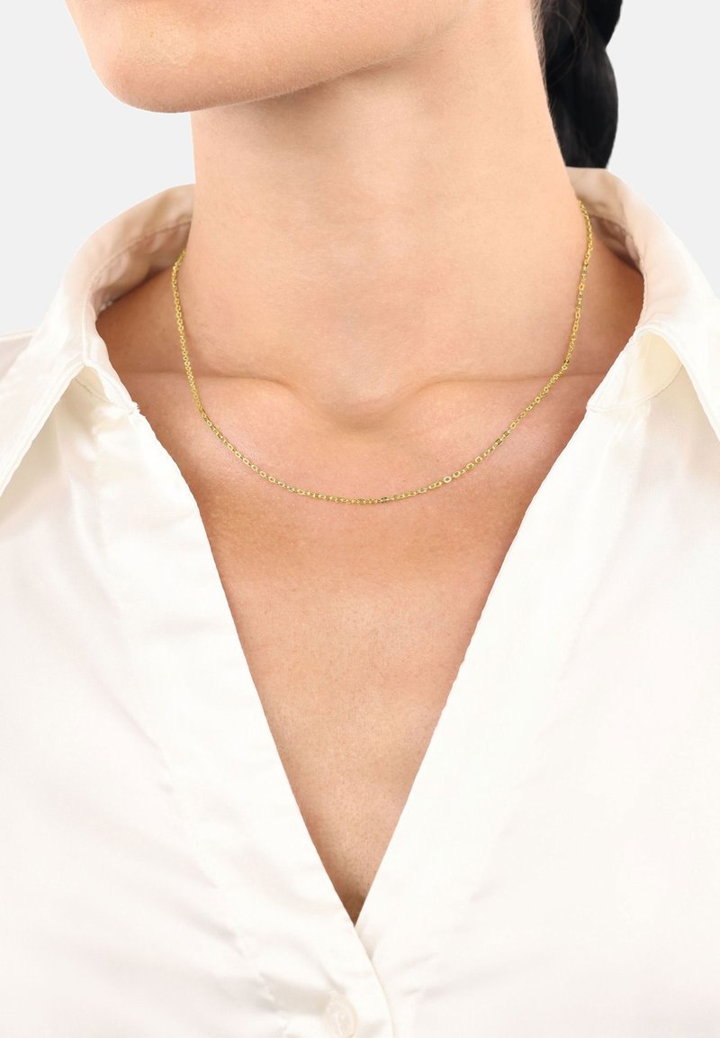 Primer plano de una persona que lleva un delicado collar de cadena de oro sobre una camisa blanca con cuello, mostrando el cuello y la parte superior del pecho.