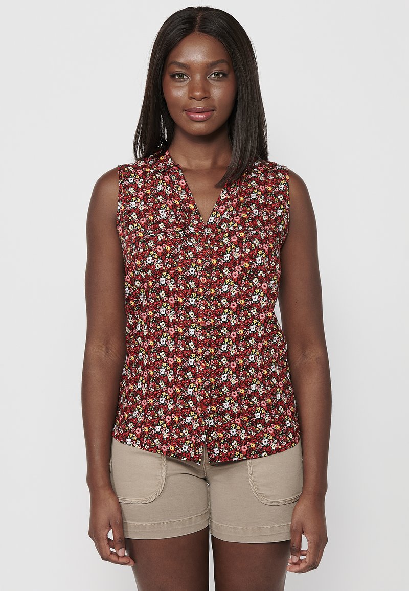 Blusa sin mangas roja de flores con escote en V, que presenta flores blancas, rosas y amarillas. Combinada con pantalones cortos beige.
