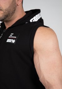 Zwart mouwloos hoodie met rits, met een textuur patch met de tekst "VOOR DE GEMOTIVEERDEN" en "GORILLA WEAR" in het wit. Gelaagde stof.