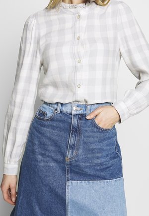 Femme portant un chemisier boutonné à carreaux blancs et gris clair avec des manches bouffantes, rentré dans une jupe en jean bleue bicolore.