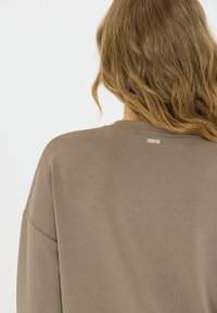 Bruine lange mouw top met ronde hals en zachte textuur. Een klein goud label bevindt zich aan de achterkant bij de halslijn.