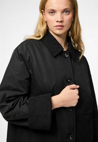 Cappotto nero con colletto in velluto a coste, grandi bottoni e vestibilità comoda. Il tessuto è liscio con una leggera lucentezza e polsini arrotolati.