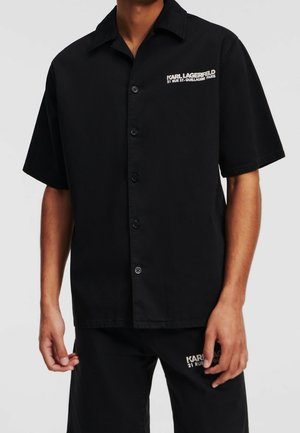 Mężczyzna ubrany w czarną, krótką koszulę na guziki i pasujące szorty z białym napisem "Karl Lagerfeld 21 Rue St-Guillaume Paris".