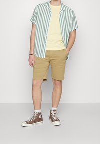 Lyhythihainen vihreä ja valkoinen raidallinen nappipaita keltaisen T-paidan päällä, yhdistettynä vaaleanbeigeihin shortseihin ja ruskeisiin nilkkureihin.