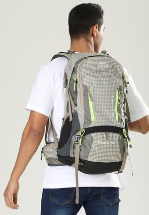 Man draagt een grijze en zwarte wandelrugzak met groene ritssluitingen, een wit shirt en een blauwe spijkerbroek, gezien van achteren.