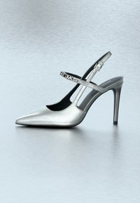 Ασημένιο παπούτσι με ψηλό τακούνι, μυτερή μύτη και ρυθμιζόμενο λουράκι με διακοσμητική πόρπη. Μεταλλική, λεία υφή με κομψό σχεδιασμό.