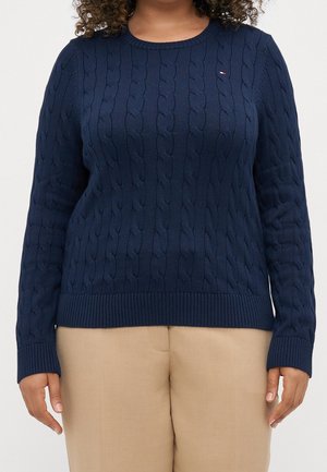 Personne portant un pull torsadé bleu marine et un pantalon beige, visible des épaules aux hanches sur un fond uni.