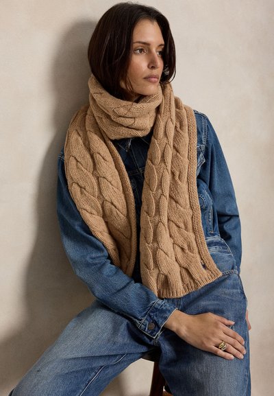 Femme assise en veste en jean bleue et jean, portant une grosse écharpe beige en maille torsadée enroulée autour du cou.