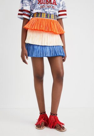 Model wearing a tiered skirt with orange, cream, and blue fringes, a colorful woven belt, and red fringed ankle accessories with sandals.