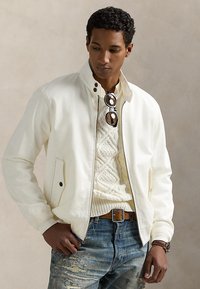 Homme portant une veste blanche zippée sur un pull à torsades, un jean bleu délavé, une ceinture marron, des lunettes de soleil suspendues au col du pull et une montre-bracelet.