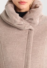 Cappotto di lana beige con colletto alto e ripiegato, caratterizzato da una texture morbida e un design minimalista. Davanti senza bottoni con dettagli a cucitura.