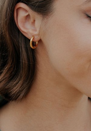 Gold hoop earring worn on the earlobe, featuring a smooth, reflective surface and a circular design. Hair is styled in a bob.