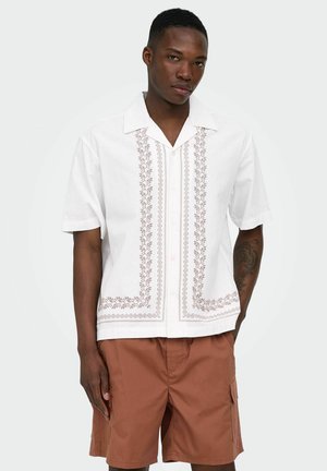 Młody mężczyzna w białej koszuli z krótkim rękawem z haftowanymi wzorami i brązowych spodenkach typu cargo, stojący na jednolitym tle.