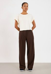 T-shirt creme cortada com mangas dobradas combinada com calças largas castanhas. Tênis pretos completam o conjunto. Design simples e cores neutras.