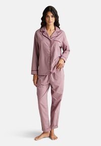 Vrouw staat blootsvoets in een roze en wit geruit pyjamastel met lange mouwen, knopenlijst, kraag en manchetten.