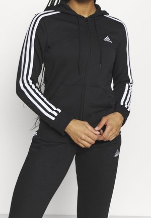 Kobieta ubrana w czarną bluzę z kapturem i zamkiem Adidas oraz pasujące spodnie z białymi paskami na rękawach i nogawkach, stojąca na białym tle.