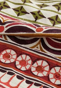 Tissu avec des motifs complexes en rouge, vert et crème. Présente diverses formes circulaires et géométriques, avec une finition texturée.