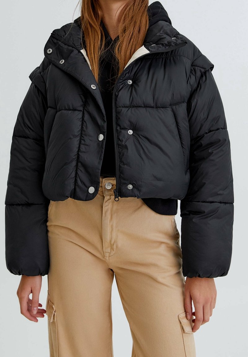 Krátká černá péřová bunda s knoflíkovým zapínáním, kapucí a prošívaným vzorem; nošena přes černý top a kombinována s béžovými kalhotami.