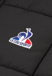 Un tissu rembourré noir présente un logo brodé avec un coq bleu, blanc et rouge, ainsi que le texte "LE COQ SPORTIF" en dessous.