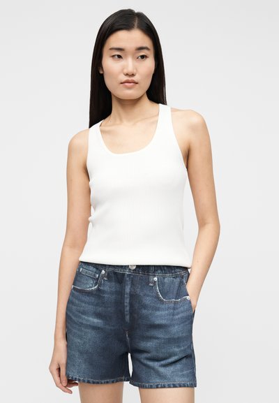 Canotta bianca a coste con spalline larghe abbinata a pantaloncini di denim scuro con tasche frontali e chiusura con bottoni, mostrata su uno sfondo uniforme.