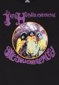 Maglietta nera con grafica iconica di una band. Immagine rotonda al centro con tre figure dai colori vivaci, circondata da scritte in viola.