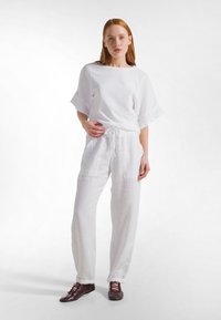 Joven mujer con cabello rojo que lleva pantalones blancos sueltos, una camiseta blanca de manga corta a juego y zapatos marrón oscuro con cordones, de pie contra un fondo blanco.