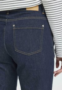 Donkerblauwe denim jeans met een klassiek ontwerp, gele stiksels, een achterzak en een leren label op de tailleband. Gestreept shirt zichtbaar erboven.