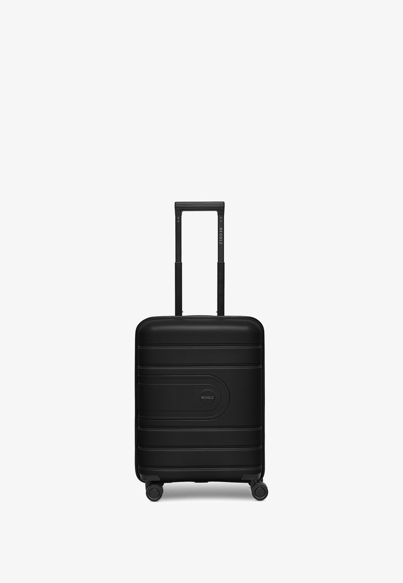 Valigia rigida nera con superficie testurizzata, dotata di manico telescopico e quattro ruote per la mobilità. Logo del marchio al centro della parte anteriore.