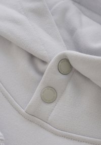 Haut gris clair avec capuche, en tissu doux, possédant un col replié et deux boutons-pression avec un texte en relief. Couture visible le long des bords.