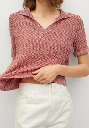 Kobieta w czerwonym, krótkim sweterku z teksturą i kołnierzykiem, podnosząca dół, by pokazać białe spodnie z wysokim stanem.