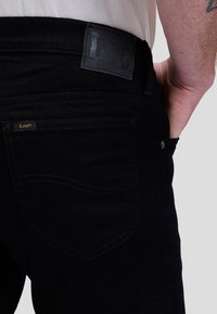 Personne portant un jean Lee noir avec la main dans la poche avant, montrant la poche arrière et le patch en cuir de la marque sur la ceinture.