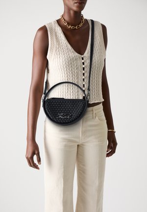 Sac à main rond noir avec une texture tissée, doté d'une fermeture éclair en argent et de détails emblématiques, porté en bandoulière sur un haut en tricot crème et un pantalon clair.