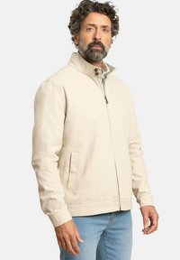 Chaqueta ligera beige con cuello alzado, cierre de cremallera y detalles de botones a presión. Cuenta con bolsillos laterales y una textura de tela suave.