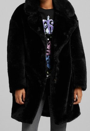 Persona che indossa un cappotto in pelliccia sintetica nera sopra una maglietta nera con grafica e jeans grigi, in piedi su uno sfondo grigio uniforme.