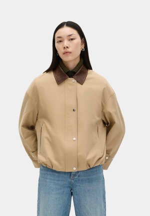 Frau mit langen schwarzen Haaren, die eine beige Jacke mit braunem Kragen, einen grünen Rollkragenpullover und blaue Jeans trägt, steht mit den Händen in den Taschen vor weißem Hintergrund.