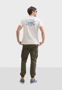 Camiseta casual de color beige claro con un gráfico azul en la parte trasera, combinada con pantalones cargo verdes oliva y zapatillas azules con acentos naranjas.