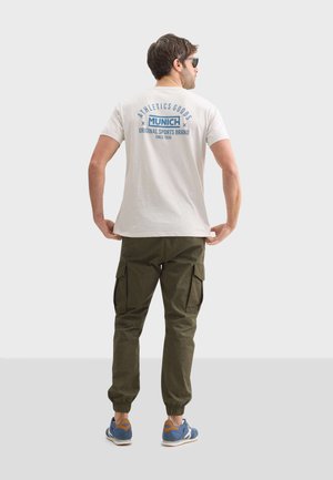 Camiseta casual de color beige claro con un gráfico azul en la parte trasera, combinada con pantalones cargo verdes oliva y zapatillas azules con acentos naranjas.