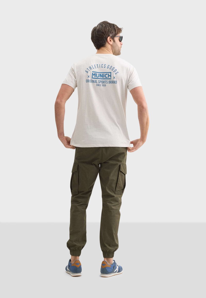 Camiseta casual de color beige claro con un gráfico azul en la parte trasera, combinada con pantalones cargo verdes oliva y zapatillas azules con acentos naranjas.