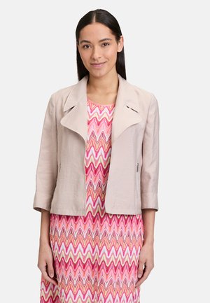Giacca attillata color beige chiaro con collo ampio, maniche corte e tasche con zip. Indossata sopra un vestito con motivo a zig-zag rosa.