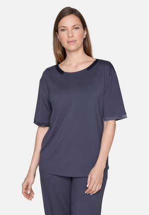 Frau mit glattem braunem Haar, die ein lockeres dunkelblaues Kurzarmshirt und passende Hose trägt, vor einem schlichten hellen Hintergrund stehend.