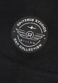 Schwarzer Stoff mit einem runden gewebten Etikett, das weißen Text, eine Flugzeuggrafik und Flügelmotive zeigt, mit der Aufschrift "Spitzbub Studios Sky Collection."