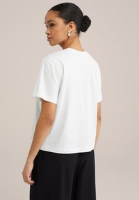 T-shirt en coton blanc à coupe décontractée, col rond et manches courtes. Porté avec un pantalon ample et sombre, mettant en valeur un design simple.