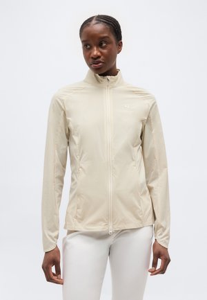 Frau mit geflochtenem Haar, die eine beige Jacke mit Reißverschluss und weiße Hose trägt, steht vor einem schlichten, hellen Hintergrund.